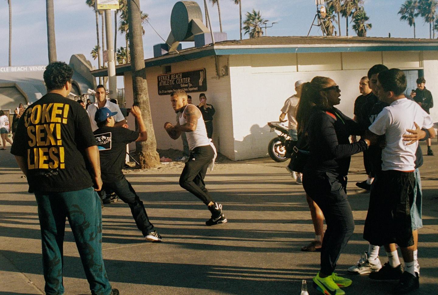 A group of people are gathered on a street with palm trees and a building in the background.