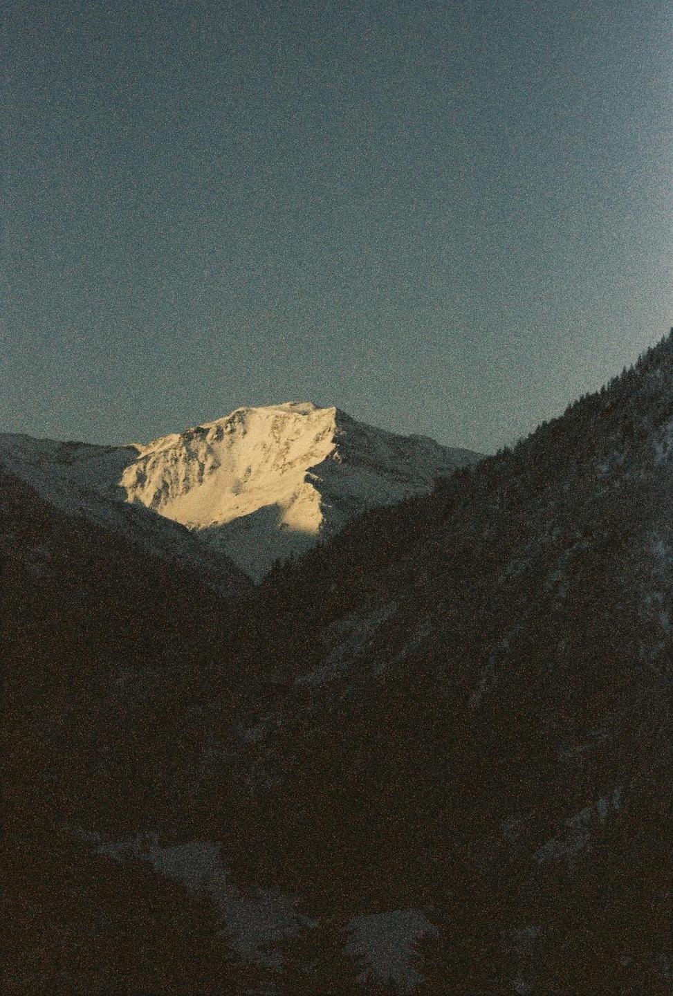 A snow-covered mountain peak is illuminated by sunlight, contrasting with the dark, shadowed mountains in the foreground.