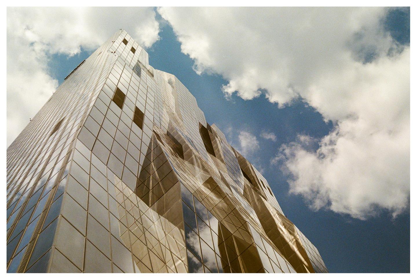 A modern glass building with a geometric facade reflects the sky and clouds.
