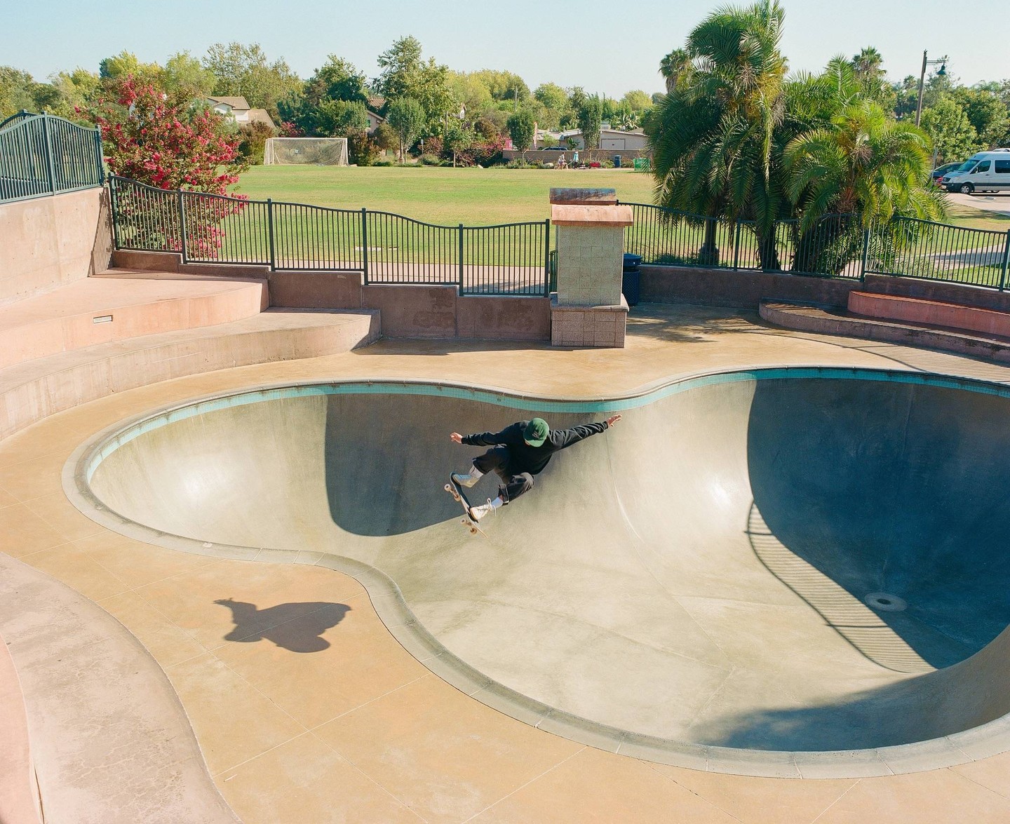 A skateboarder rides in a concrete skatepark bowl next to a grassy field with trees.