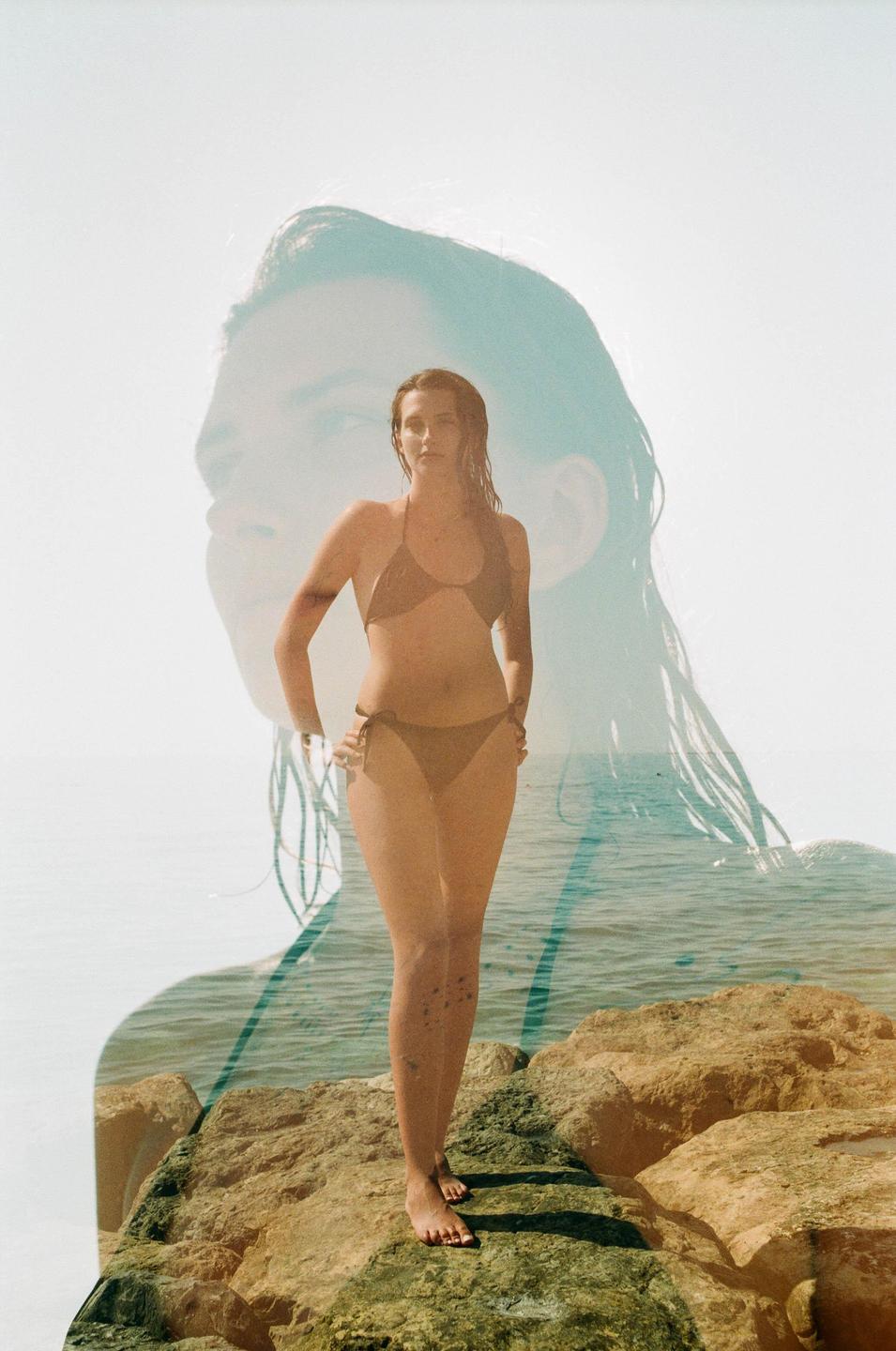 A woman in a bikini stands on rocks by the ocean with a faint double exposure of a woman's face in the background.