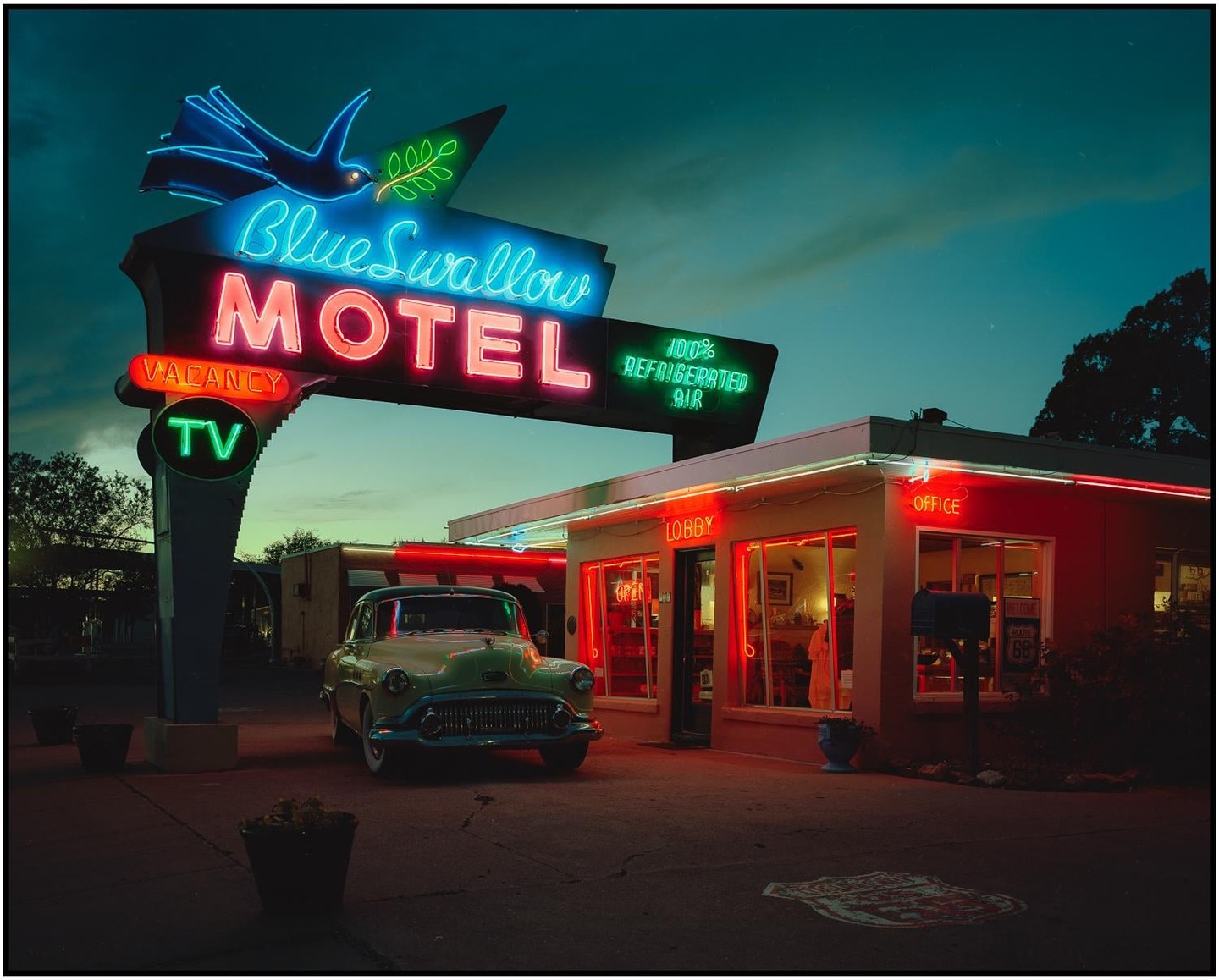 A vintage car is parked in front of the Blue Swallow Motel, which is illuminated by neon signs at dusk.