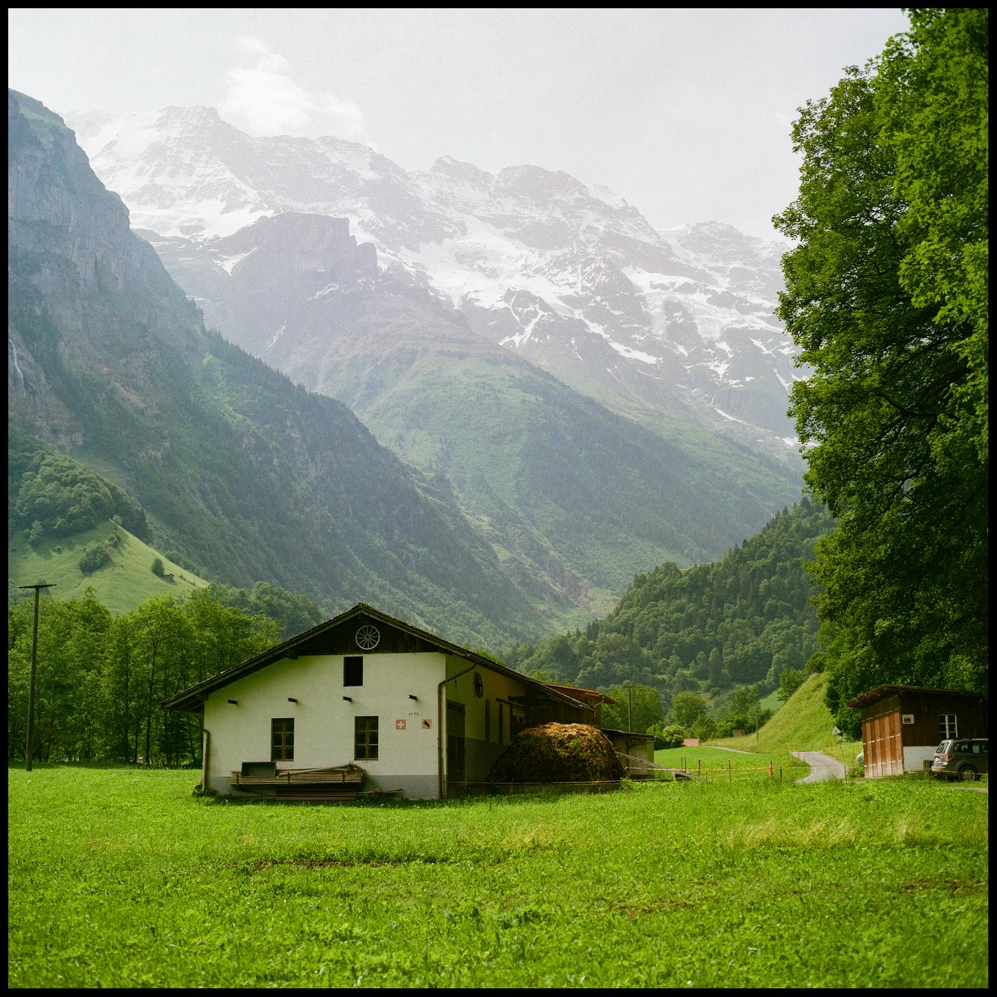 A white house with a hay bale sits in a green field with snow-capped mountains in the background.