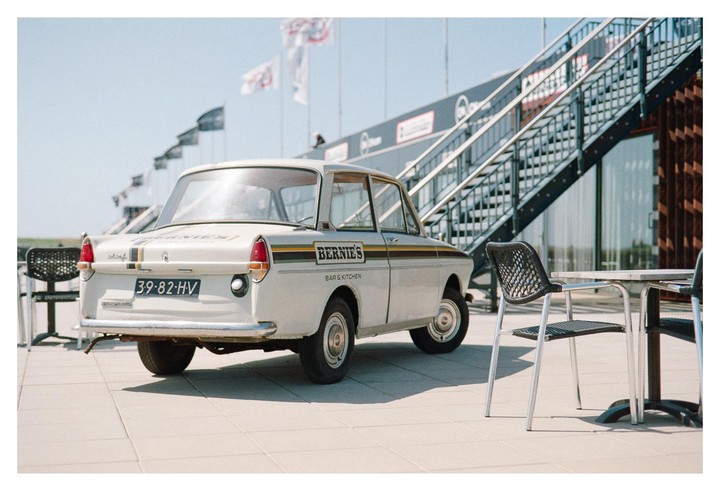 A white car with "Bernie's Bar & Kitchen" written on its side is parked next to a table and chairs, with a metal staircase and flags in the background.