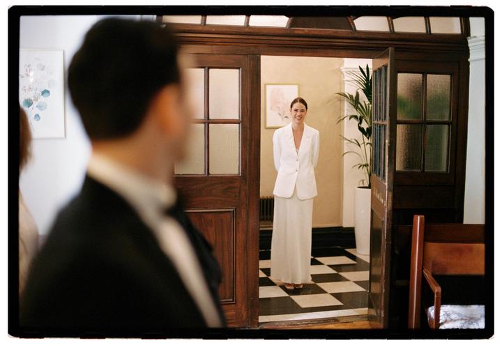A woman in a white suit smiles while standing in a doorway with a man in a suit in the foreground.