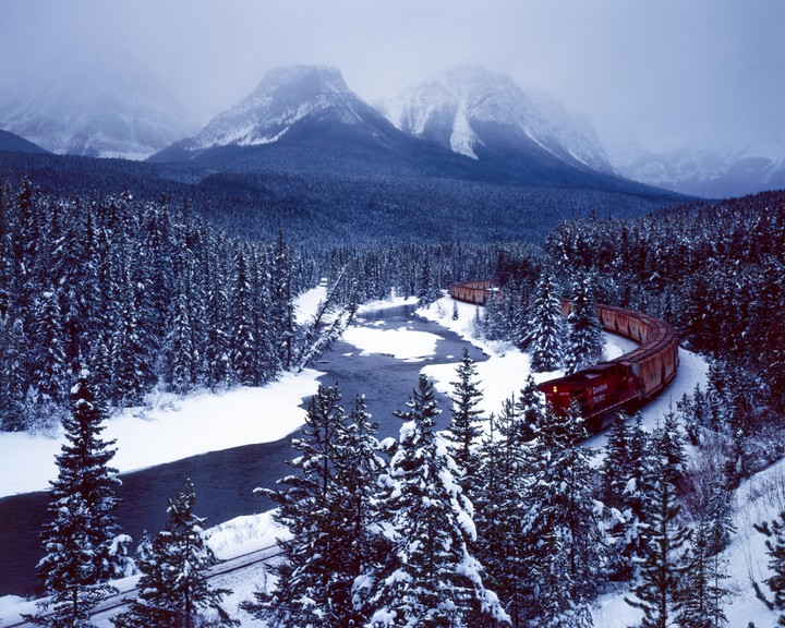 A long red train curves along a snowy riverbank surrounded by snow-covered pine trees and mountains.