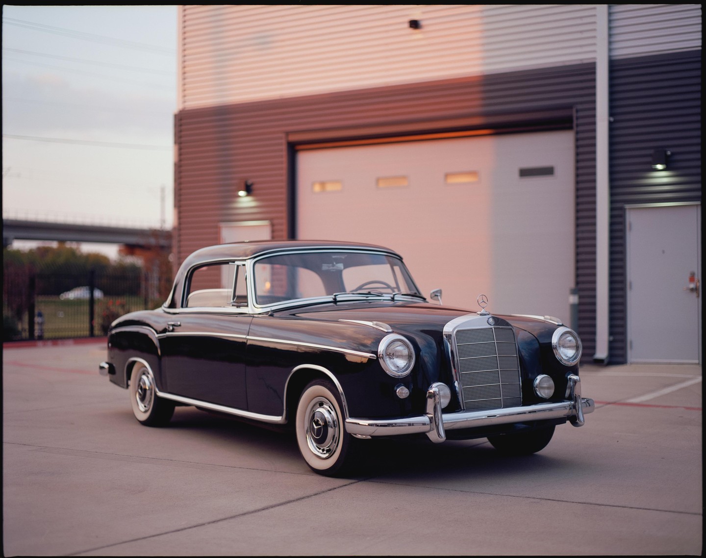 A black vintage Mercedes-Benz 220 SE is parked on a paved lot in front of a building with garage doors.