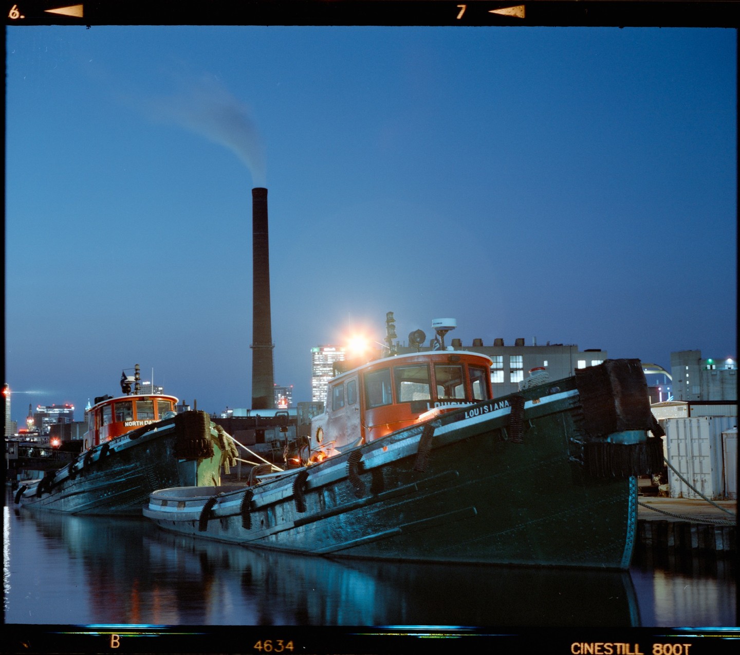 Two tugboats are docked in the water at night with a tall smokestack emitting smoke in the background.