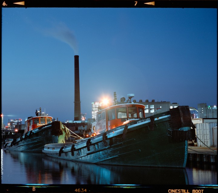 Two tugboats are docked in the water at night with a tall smokestack emitting smoke in the background.
