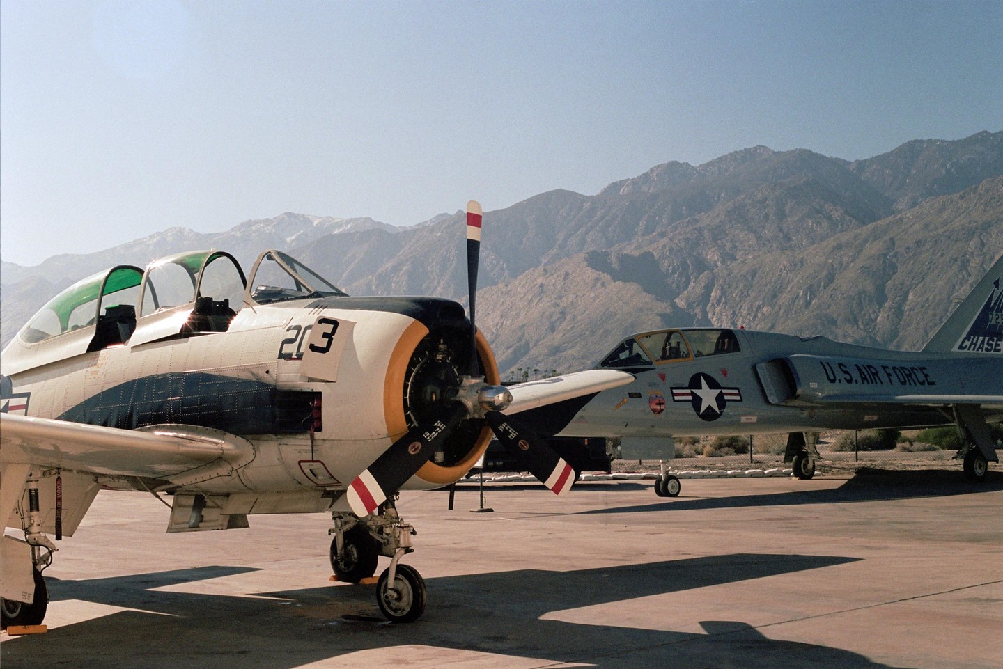 Two military aircraft are parked on an airfield with mountains in the background.