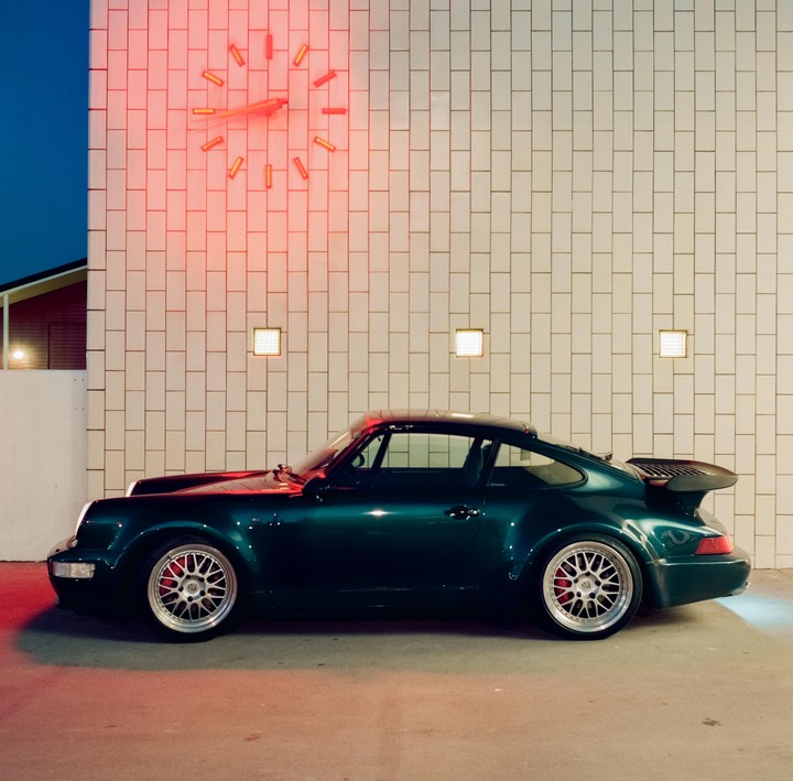 A dark green sports car is parked next to a white tiled wall with a red neon clock.