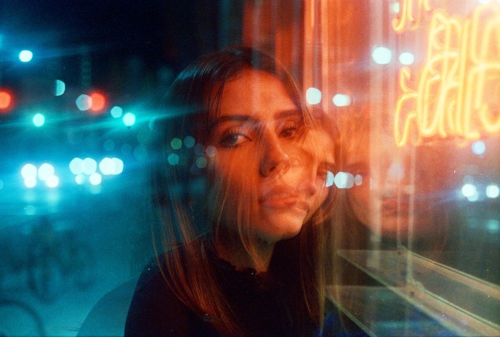 A woman's face is visible through a window with neon lights and streetlights creating a double exposure effect.