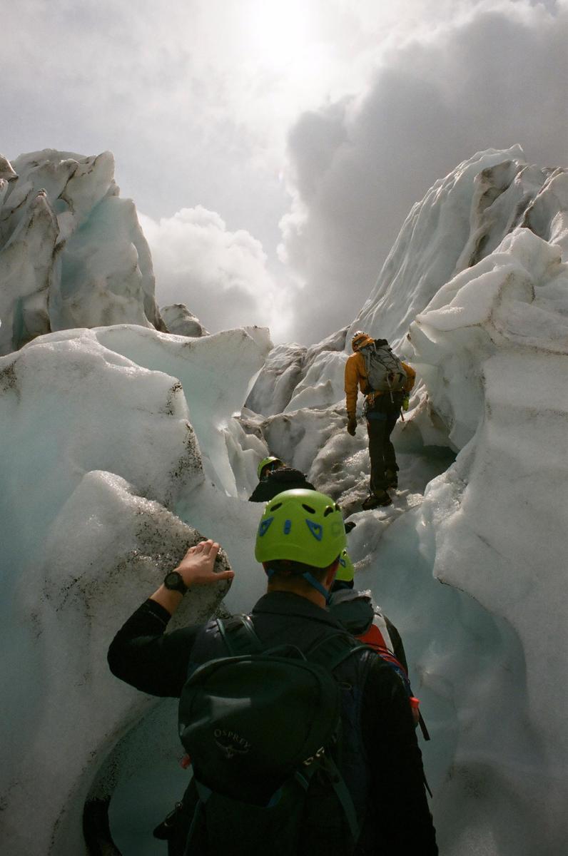 Three people wearing helmets and backpacks hike through a glacier under a cloudy sky.