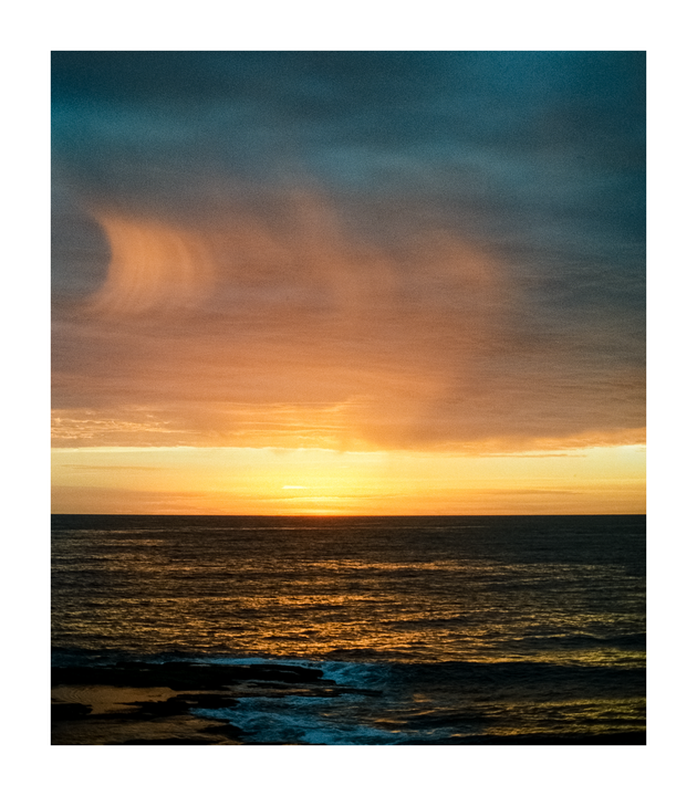 The sun sets over the ocean, casting an orange glow on the water and clouds.