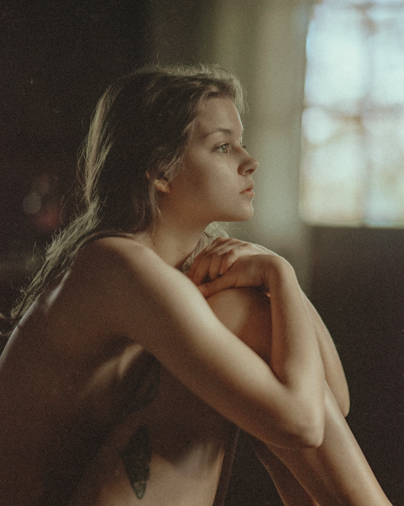 A woman with long hair sits with her arms wrapped around her knees, looking out of frame.