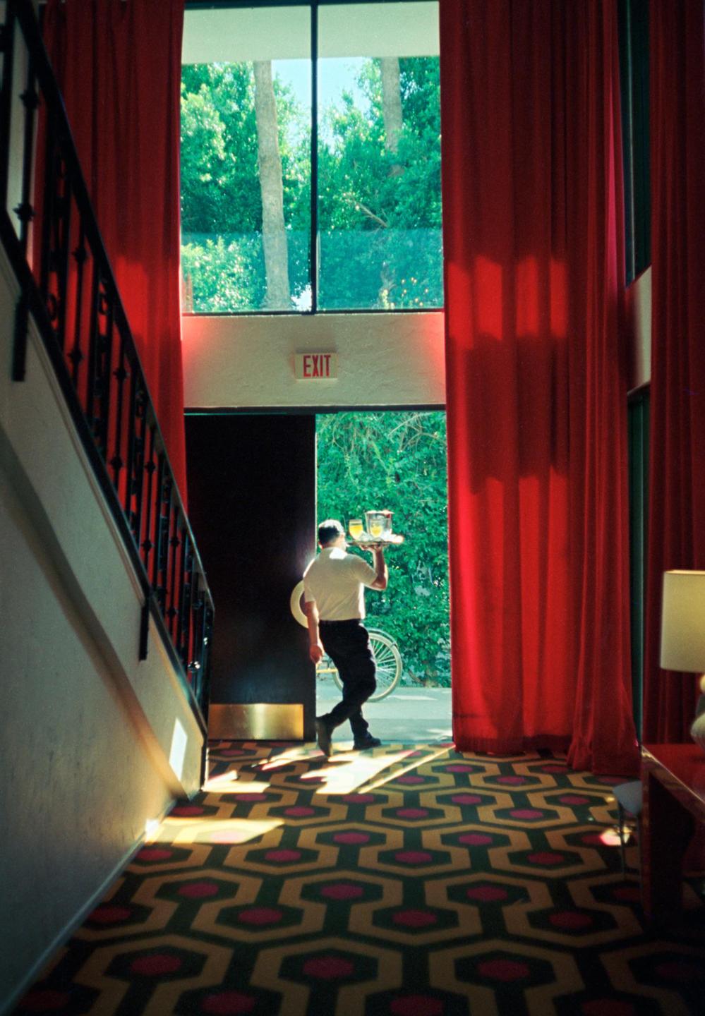 A man carrying a tray of drinks walks through a doorway with red curtains and a patterned carpet.