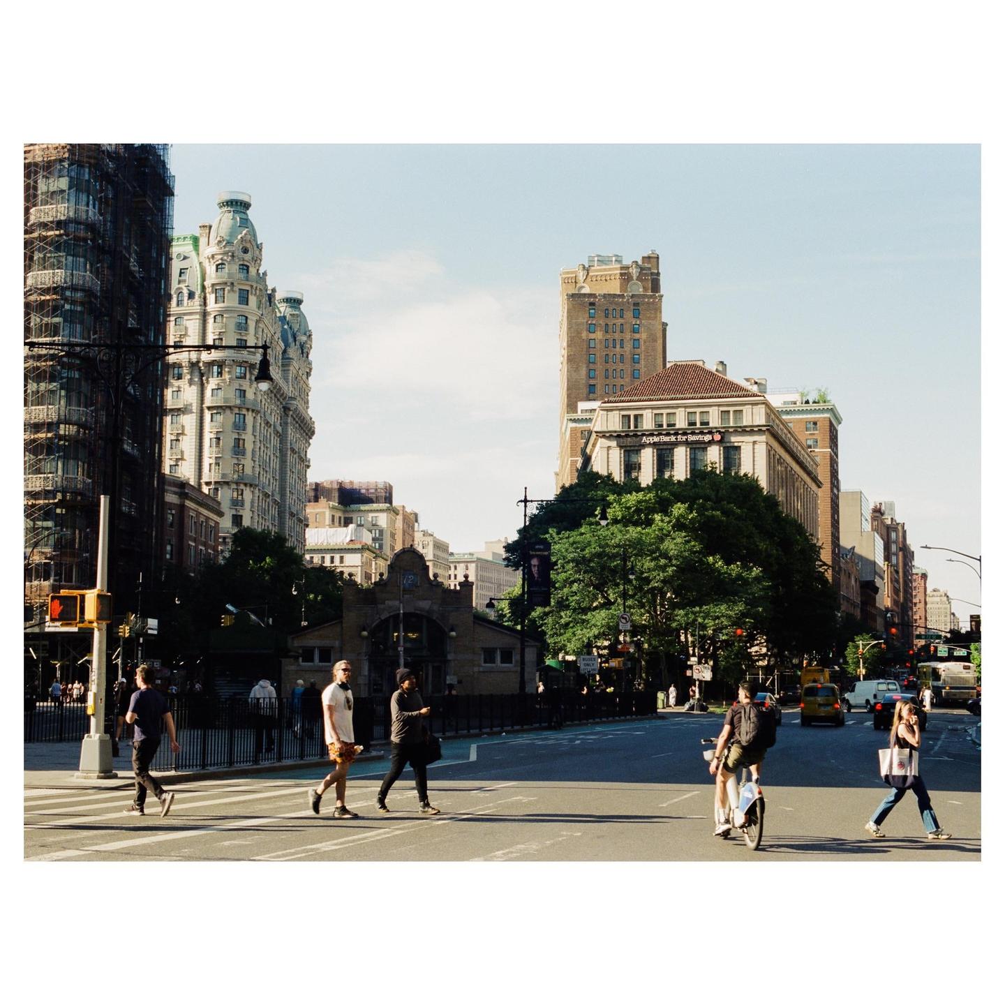 People cross a street in a city with tall buildings and trees.