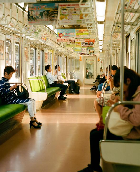 People are sitting on green seats inside a train car with advertisements hanging from the ceiling.