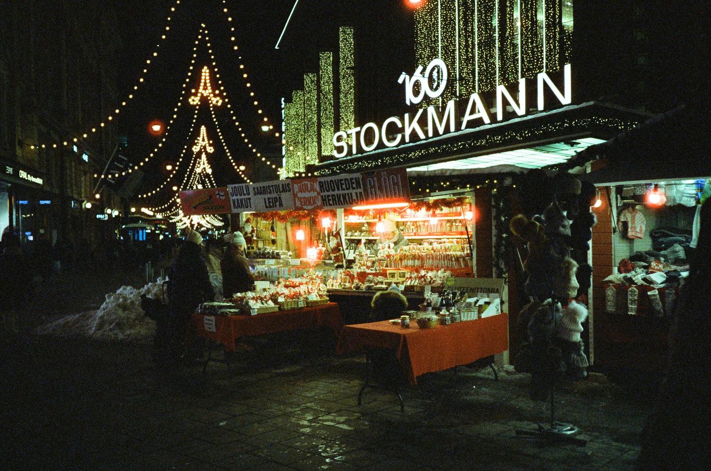 A Christmas market at night features stalls with goods and a large "Stockmann" sign illuminated by festive lights.