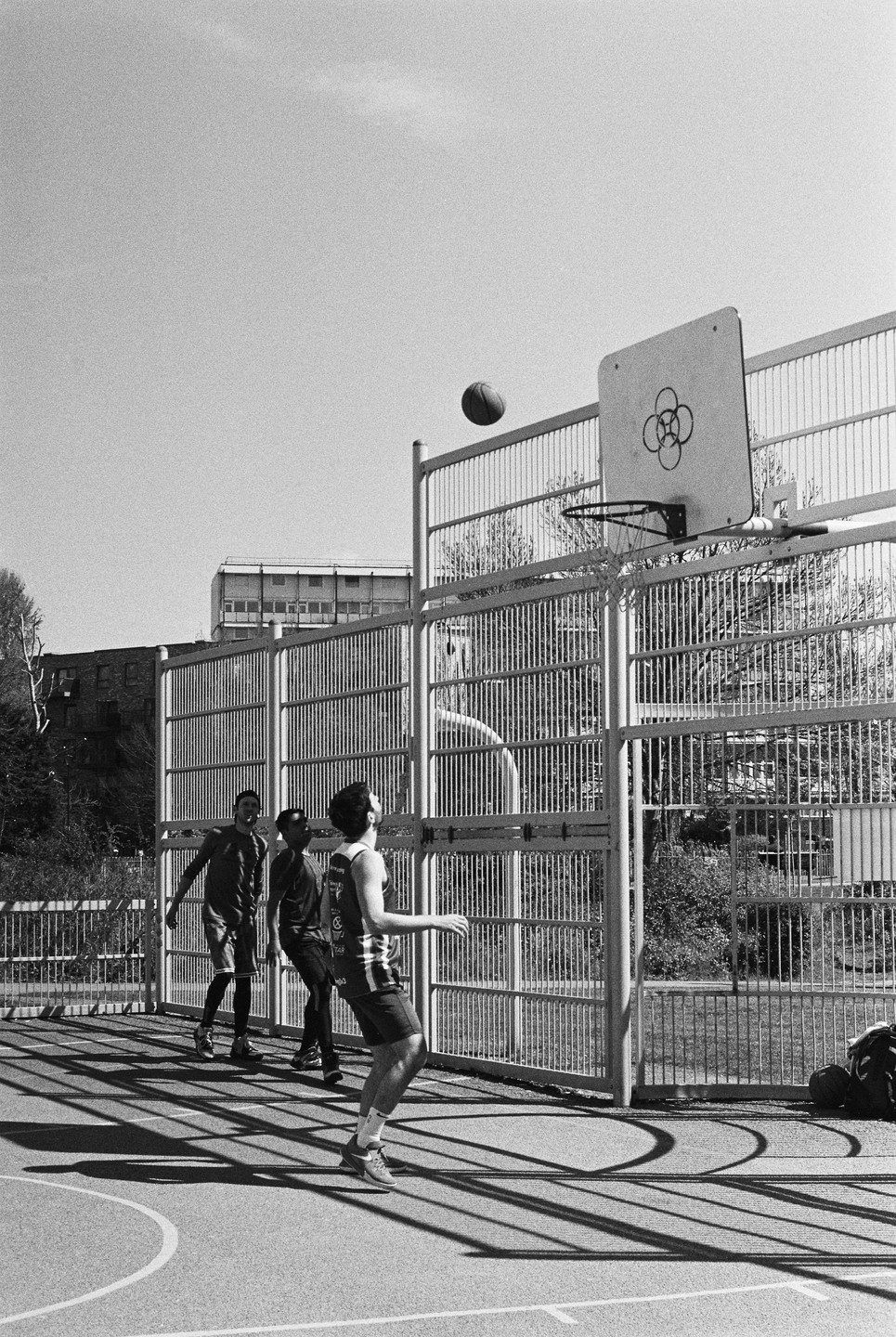Three men play basketball on an outdoor court with a ball in mid-air near the hoop.