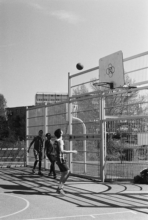 Three men play basketball on an outdoor court with a ball in mid-air near the hoop.