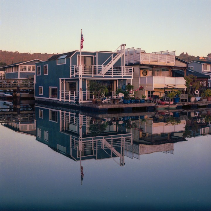 A blue houseboat with an American flag is reflected in the still water at dawn.