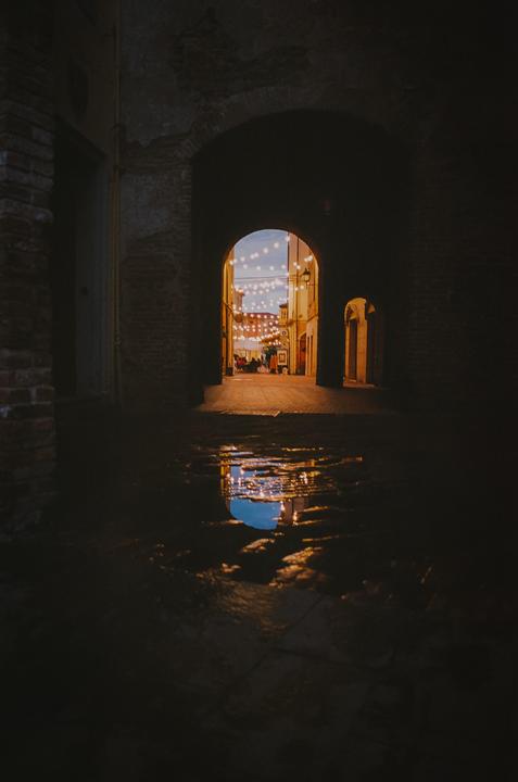 A puddle reflects string lights and a blue sky through a dark archway in an Italian city.