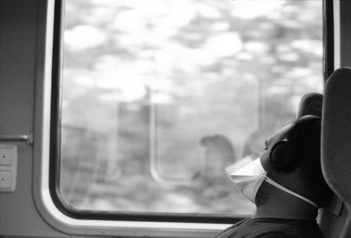 A person wearing a face mask and headphones looks out a train window.