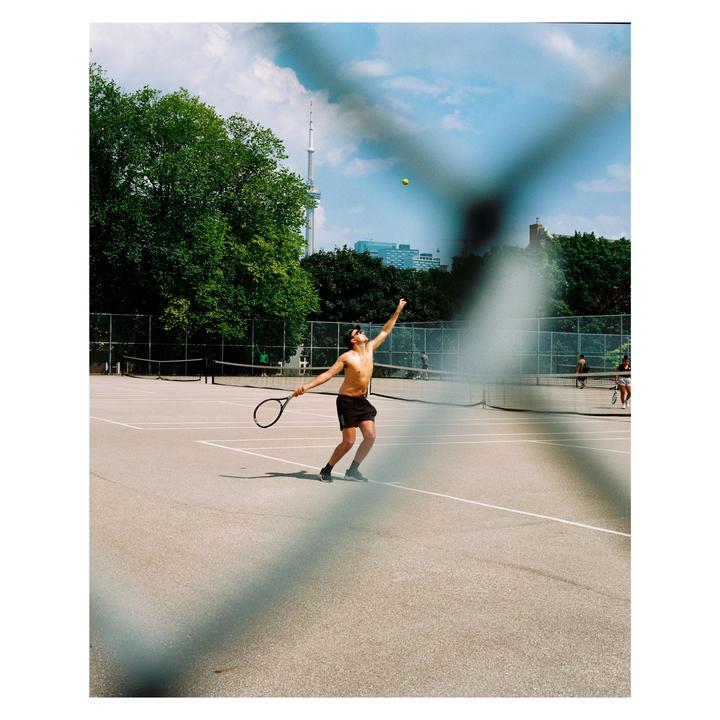 A shirtless man serves a tennis ball on a court with a city skyline in the background.