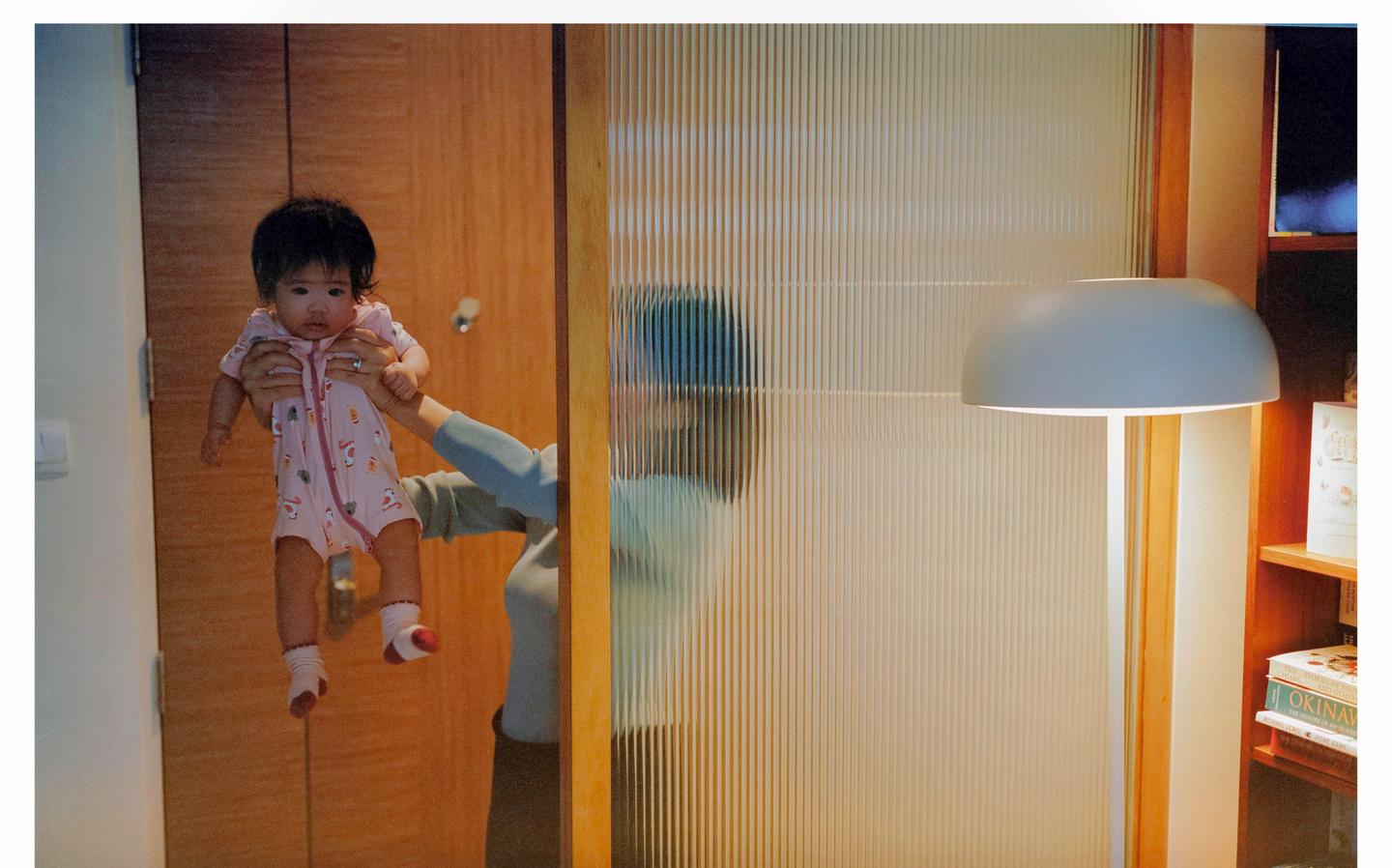 A baby in a pink onesie is held up by a woman whose reflection is visible in a fluted glass door.