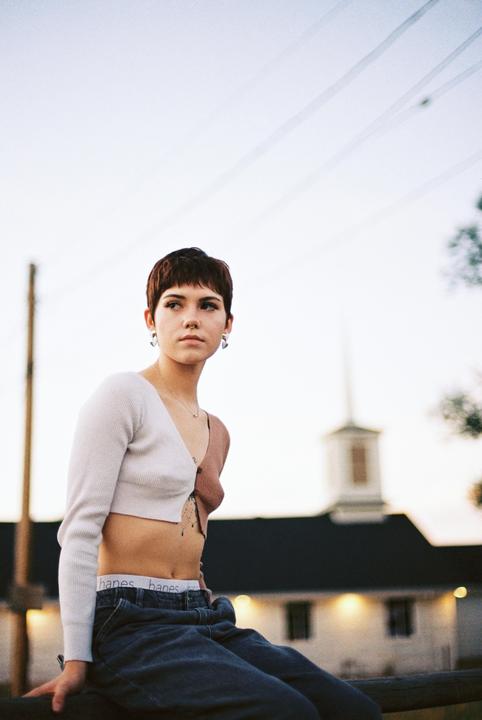 A woman with short hair and a crop top sits outdoors with a building and power lines in the background.