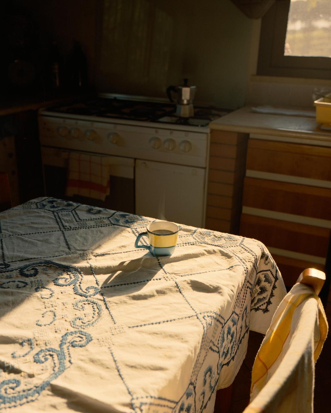 A kitchen table with a patterned tablecloth and a mug is illuminated by a shaft of sunlight.