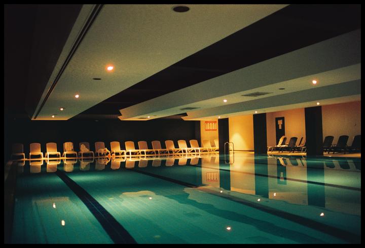 An empty indoor swimming pool with lounge chairs lining the edges is illuminated by overhead lights.