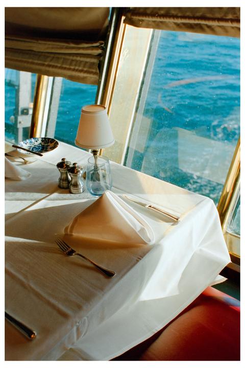 A set table with a white tablecloth, silverware, and a small lamp is next to a window overlooking the ocean.