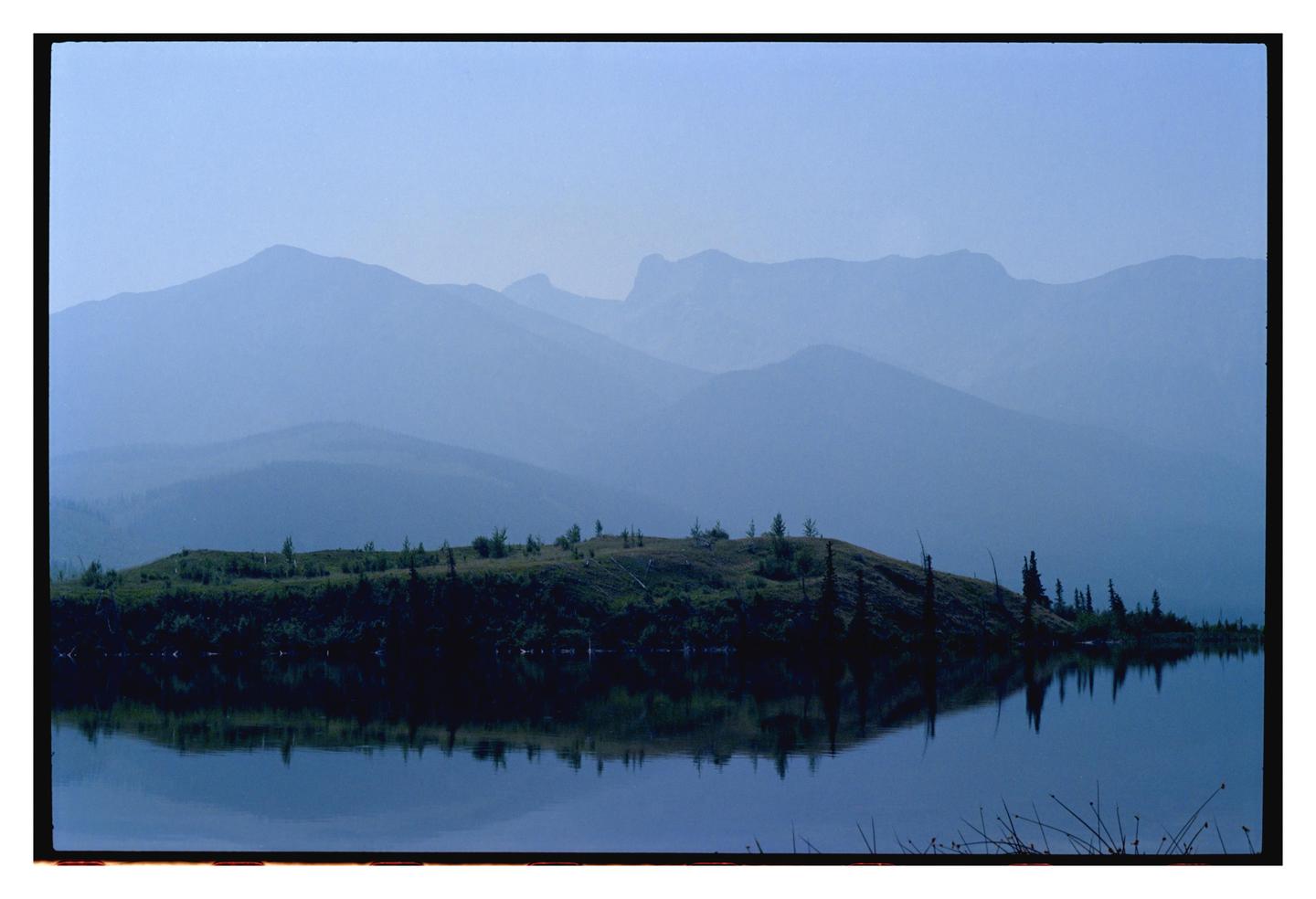 A hazy blue landscape shows a lake reflecting a tree-covered hill, with layers of mountains in the background.