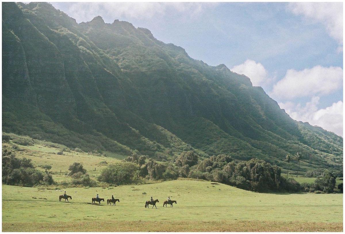 Four people ride horses across a green field with a large mountain in the background.