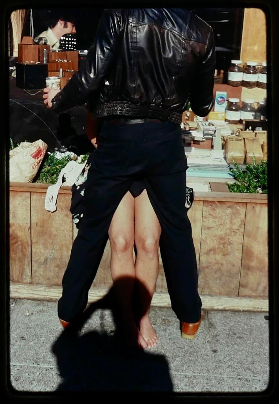 A man in a leather jacket stands in front of a stall with a barefoot woman between his legs.