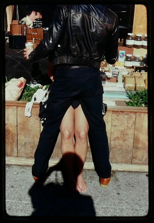 A man in a leather jacket stands in front of a stall with a barefoot woman between his legs.
