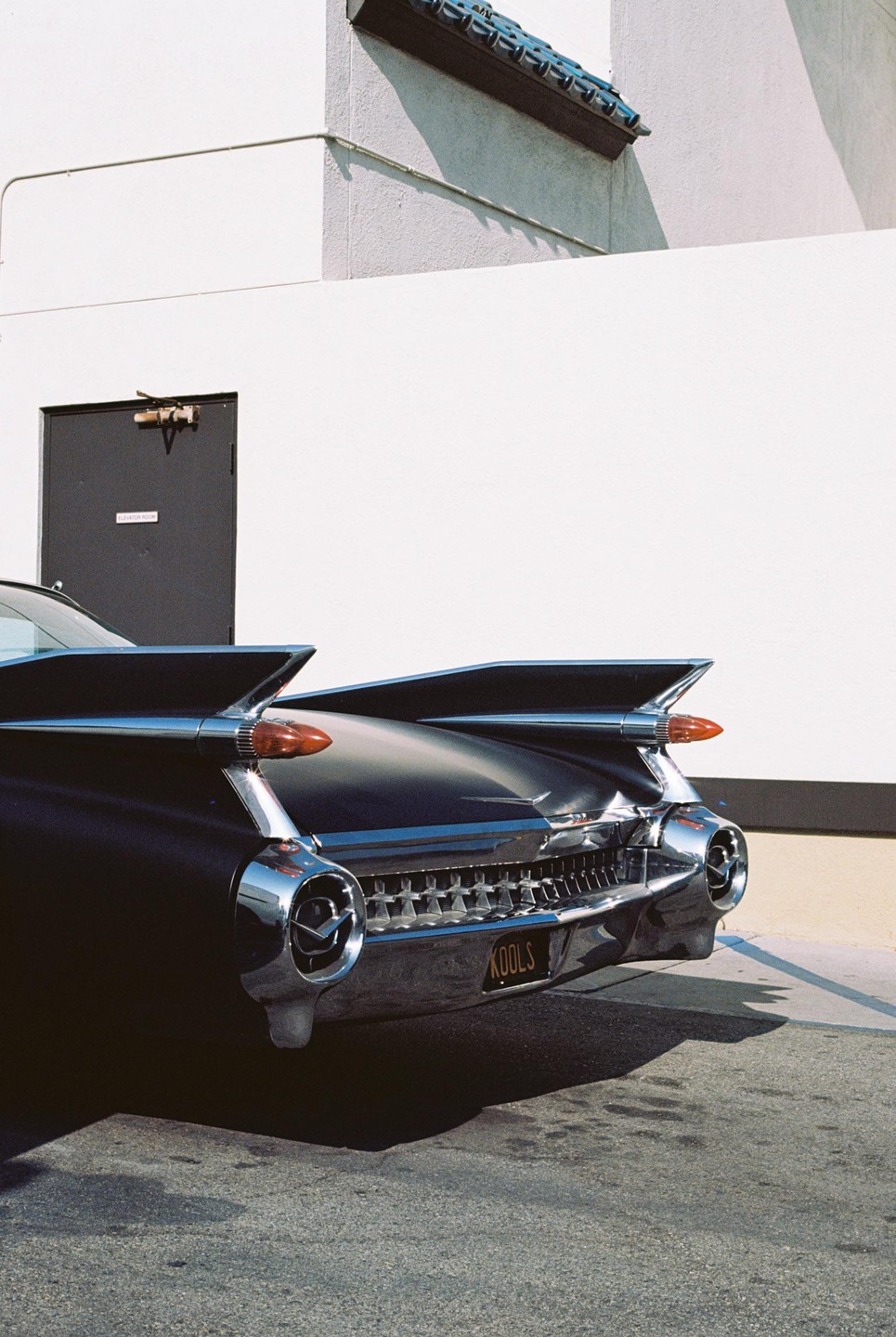 The rear of a black vintage car with prominent tail fins is parked next to a white building with a dark door.