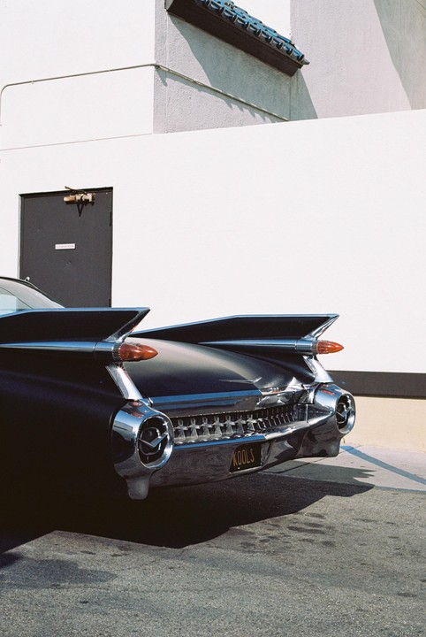 The rear of a black vintage car with prominent tail fins is parked next to a white building with a dark door.