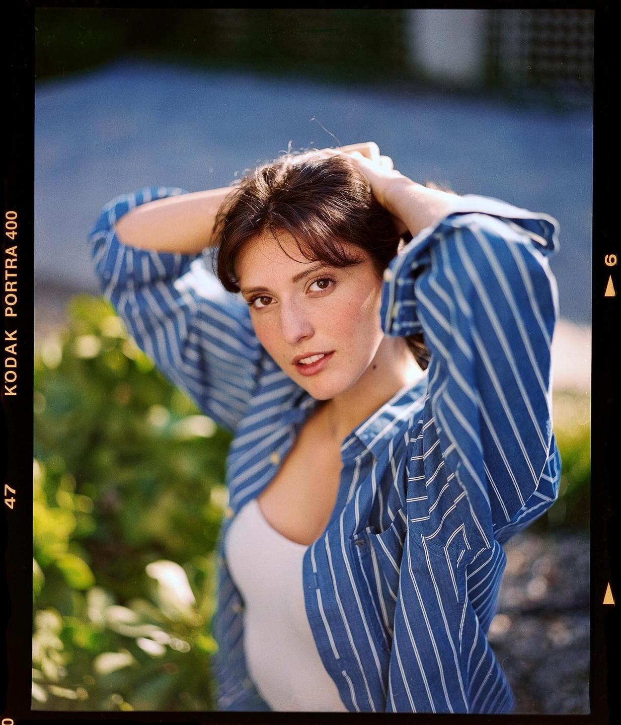 A woman with brown hair and a blue striped shirt poses with her hands behind her head.