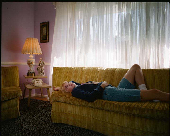 A woman with blonde hair lies on a yellow couch in a room with lavender walls and sheer white curtains.