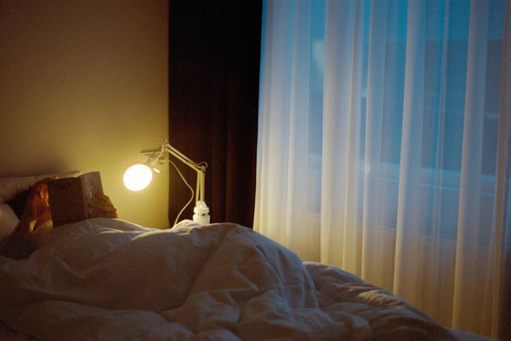 A person reads a book in bed under the light of a lamp, with a window covered by sheer curtains in the background.