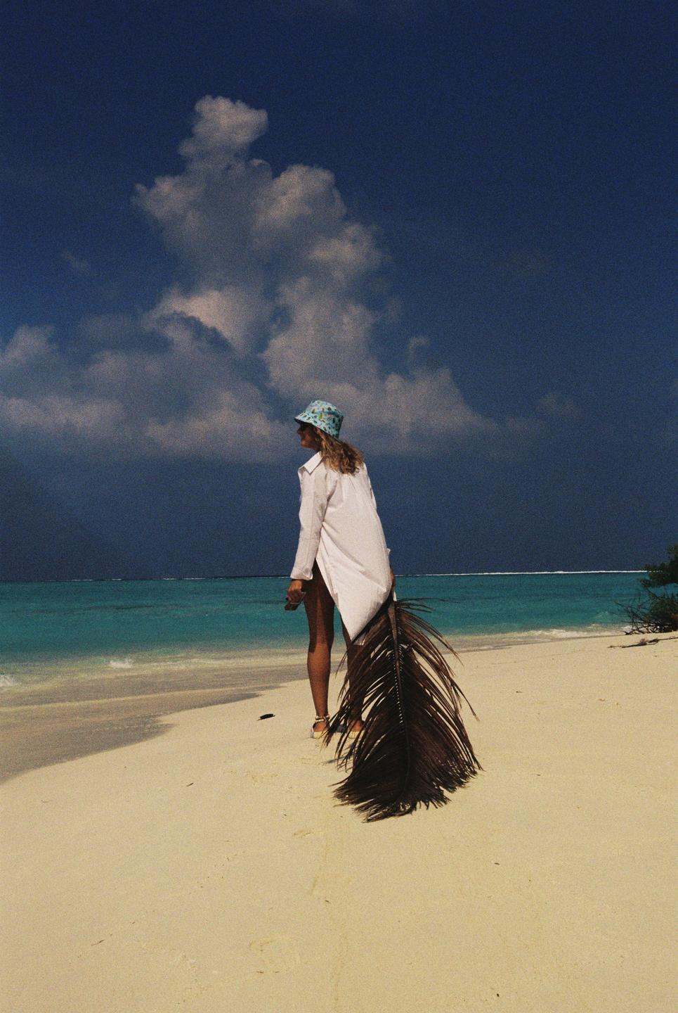 A woman in a white shirt and bucket hat stands on a sandy beach next to turquoise water, holding a large palm frond.