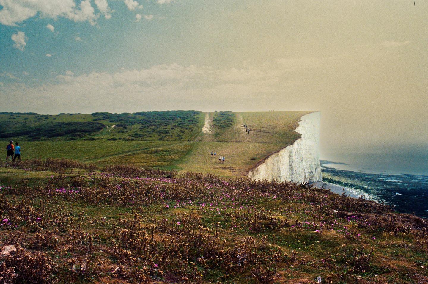 A group of people walk along a grassy cliff edge overlooking the sea under a partly cloudy sky.