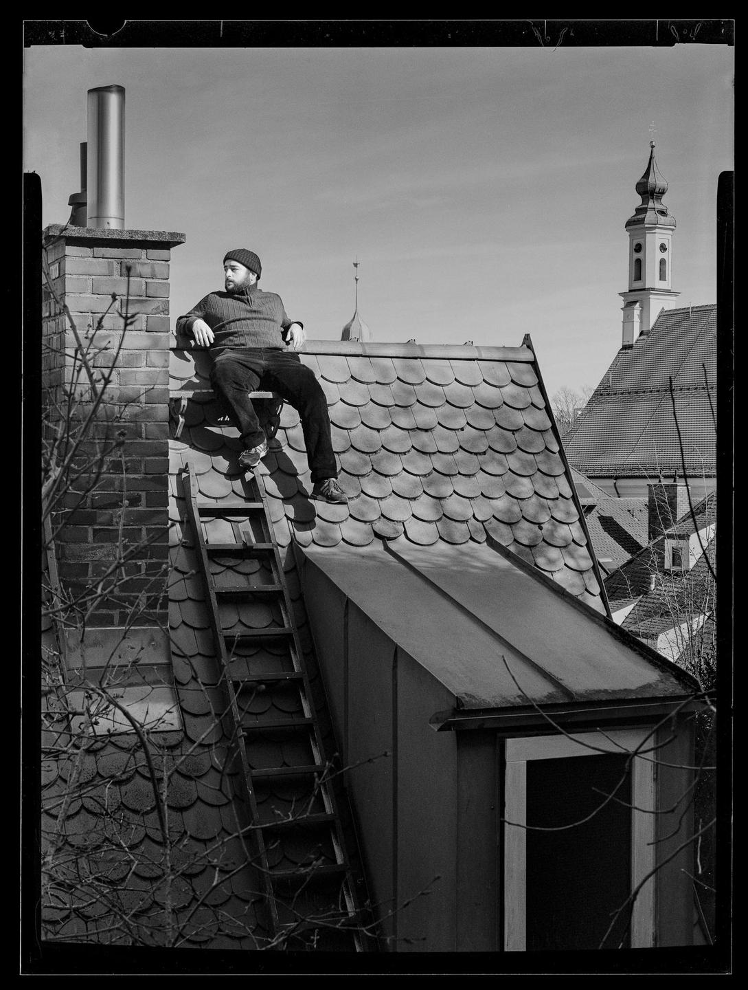 A man sits on a ladder on a tiled roof next to a brick chimney, with a church steeple visible in the background.