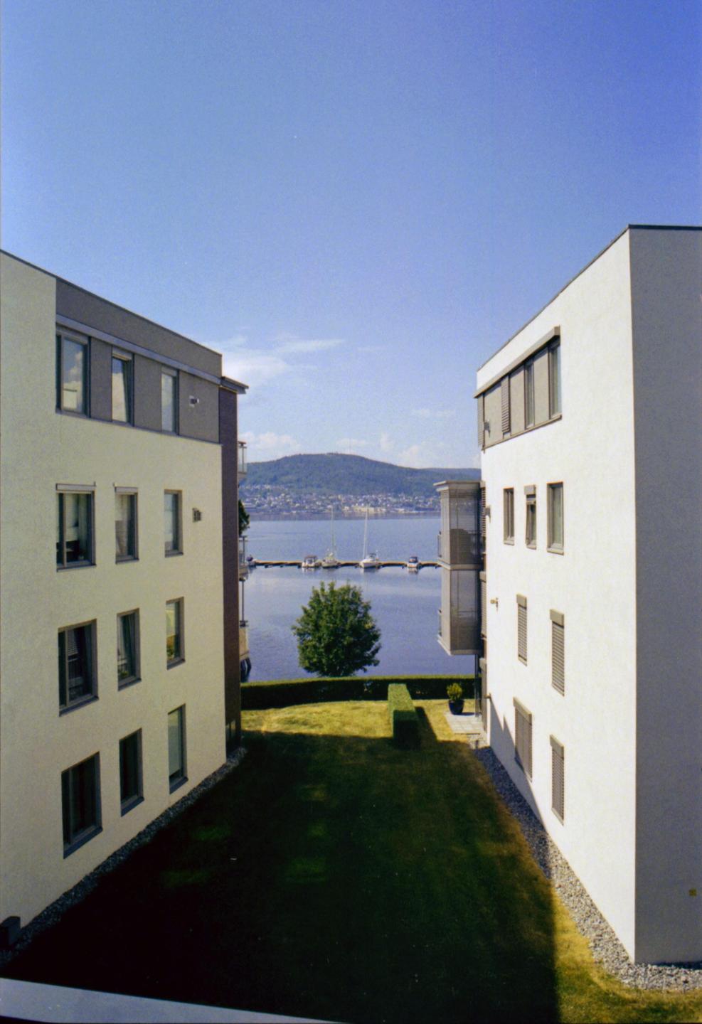 Two modern buildings flank a grassy area that leads to a body of water with boats and a distant mountain.