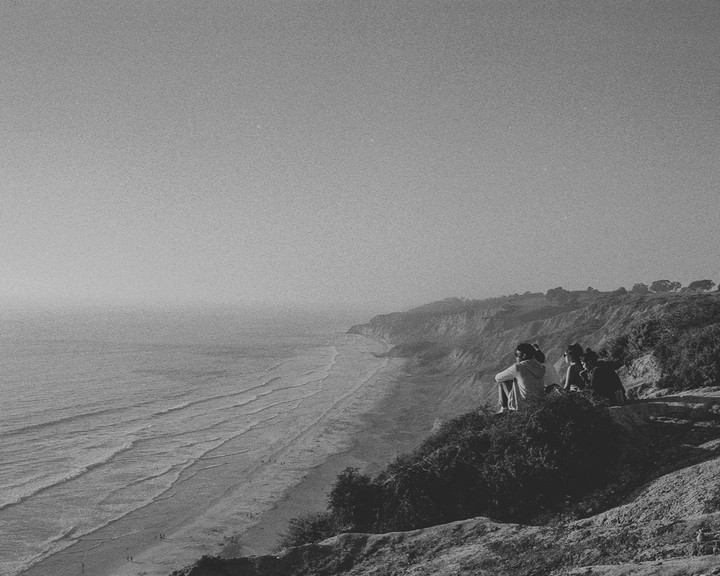 Three people sit on a cliff overlooking a beach and ocean.