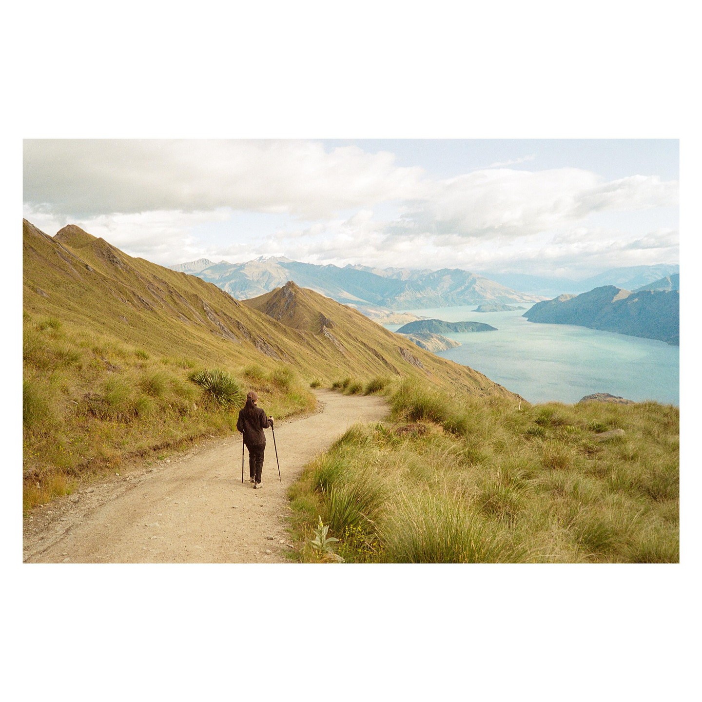 A woman walks along a dirt path on a grassy mountain overlooking a lake and distant mountains.