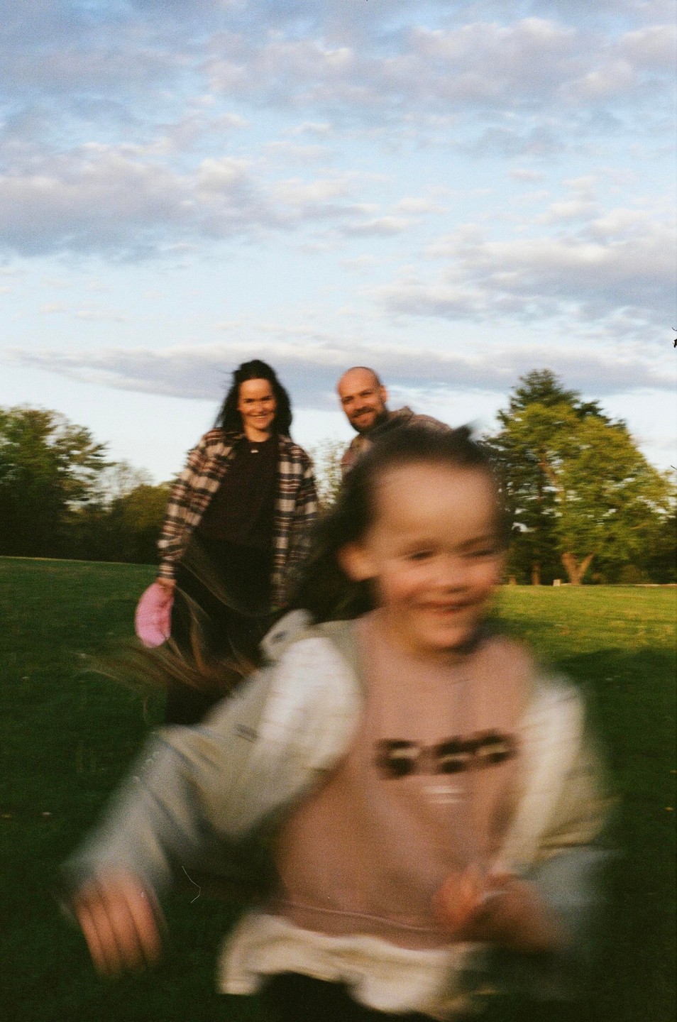 A blurred child runs towards the camera with a smiling woman and man in the background.